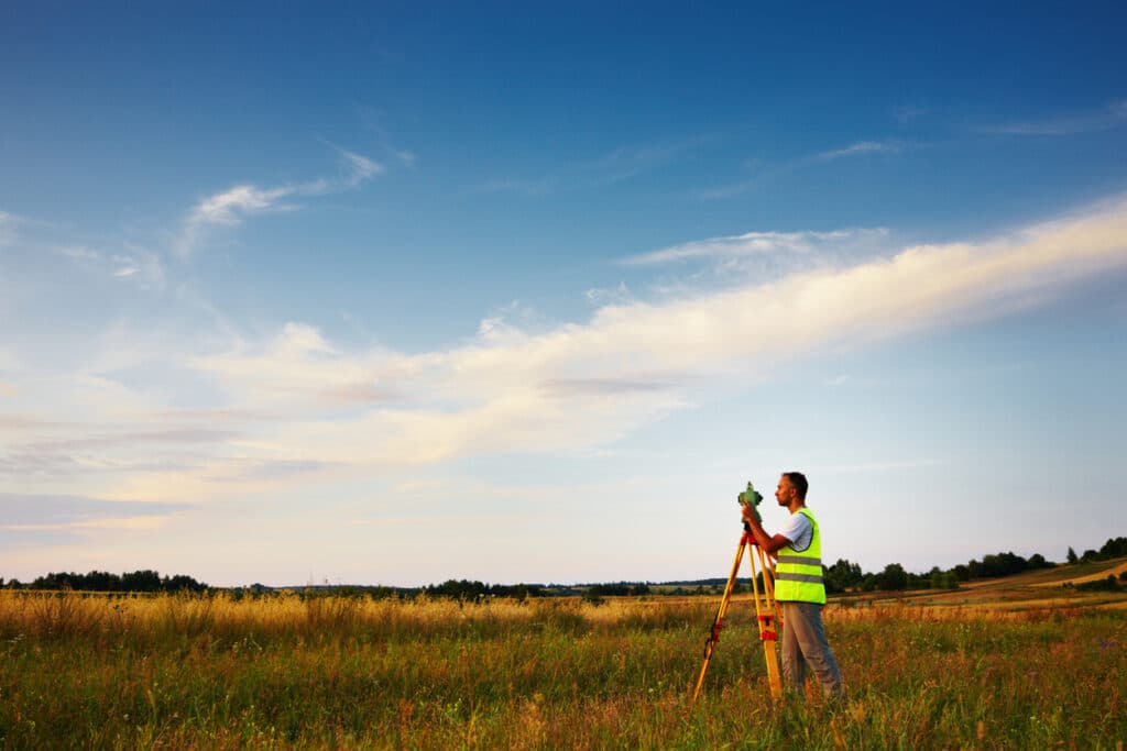 A telecommunications land surveyor wearing a high-visibility neon vest peers through his surveying instrument. In the distance is a blue sky with clouds, and a mostly flat horizon with a few trees in the background.